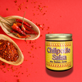 A jar of Chilpotle Salsa by Salsa Alex on a bright red background, styled with wooden spoons holding red chili peppers and chili powder.