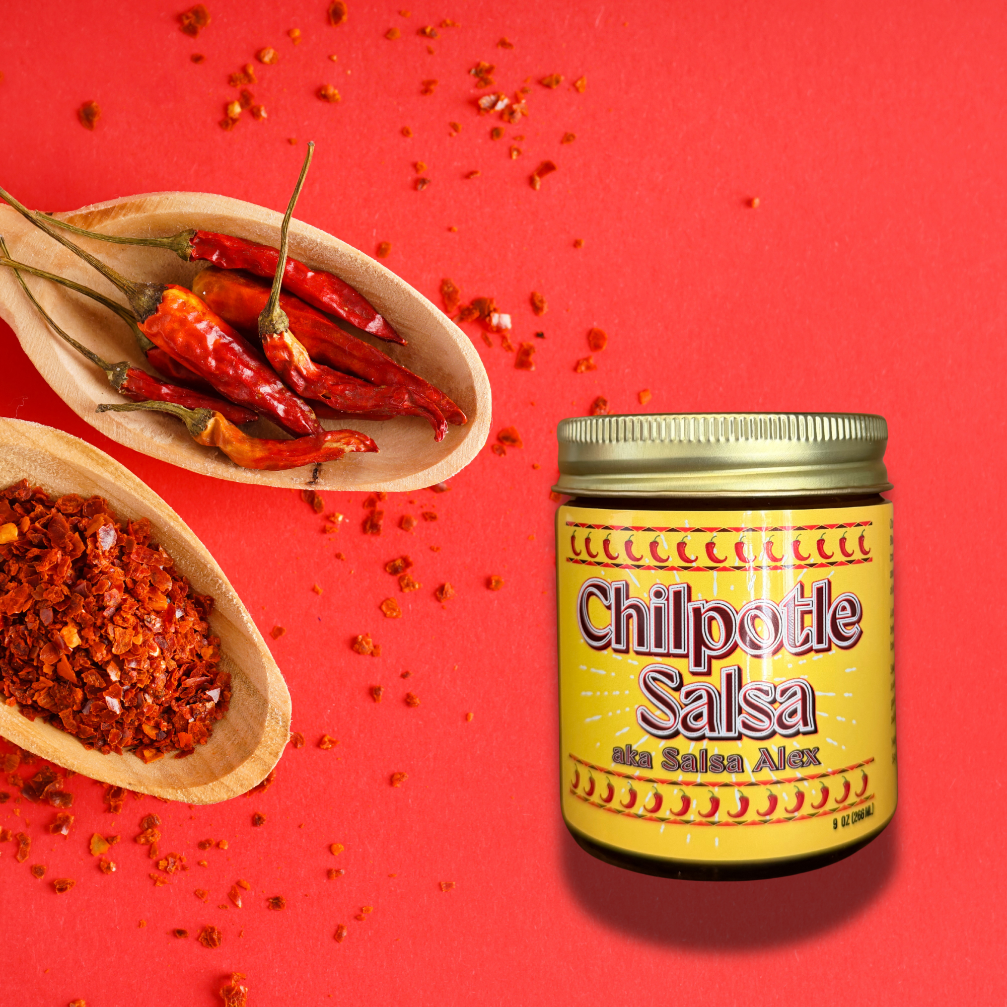 A jar of Chilpotle Salsa by Salsa Alex on a bright red background, styled with wooden spoons holding red chili peppers and chili powder.